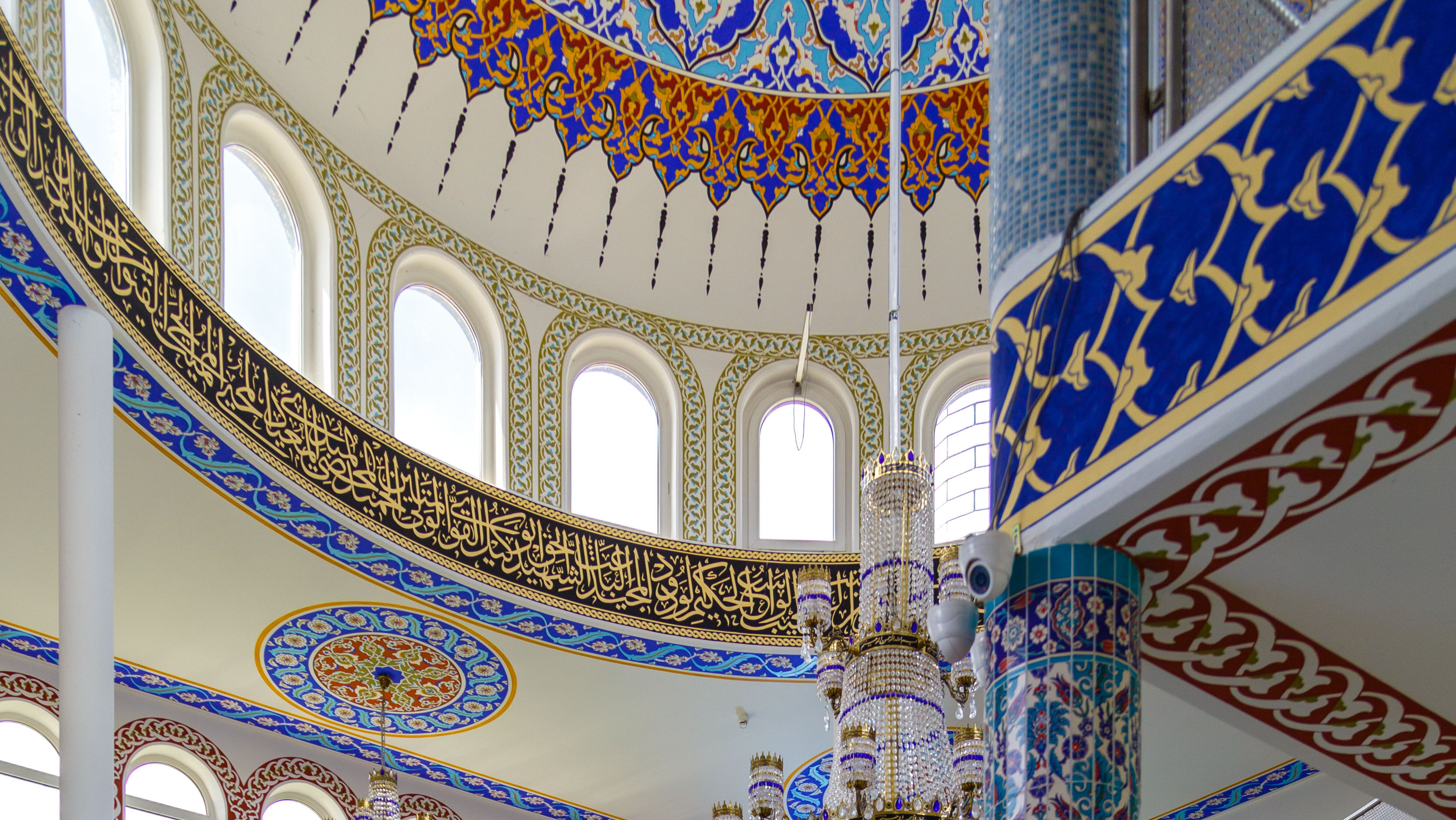 Foto des Gebetsraums für Männer in der Emir Sultan Moschee, Darmstadt. An den Wänden befinden sich bunte, verzierte Fliesen, auf dem Boden ist ein ebenso detailierter Teppich und in der Mitte des Raums hängt ein riesiger Kronleuchter mit goldenem Gestell, behangen mit einer Vielzahl an Kristallglas