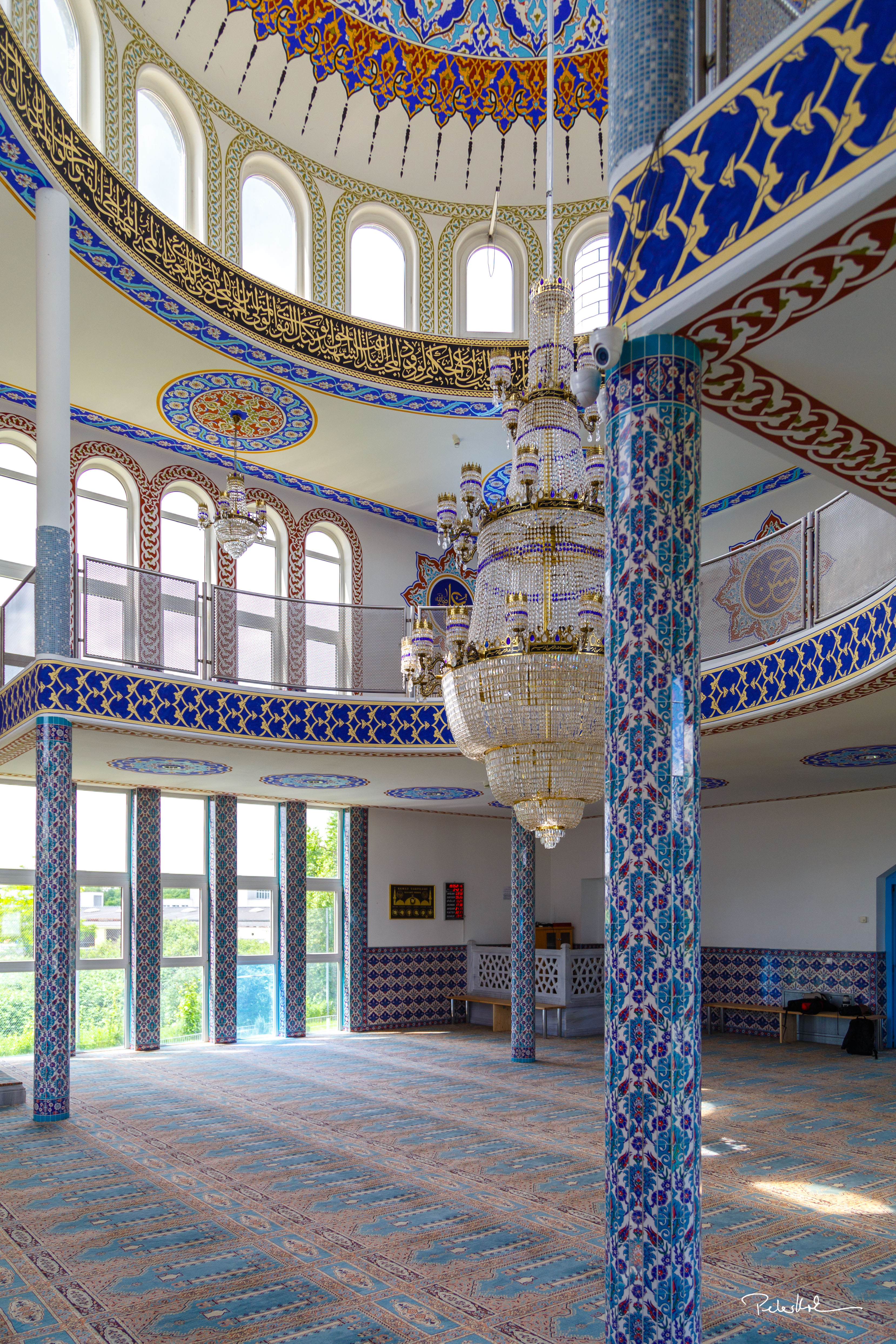 Foto des Gebetsraums für Männer in der Emir Sultan Moschee, Darmstadt. An den Wänden befinden sich bunte, verzierte Fliesen, auf dem Boden ist ein ebenso detailierter Teppich und in der Mitte des Raums hängt ein riesiger Kronleuchter mit goldenem Gestell, behangen mit einer Vielzahl an Kristallglas