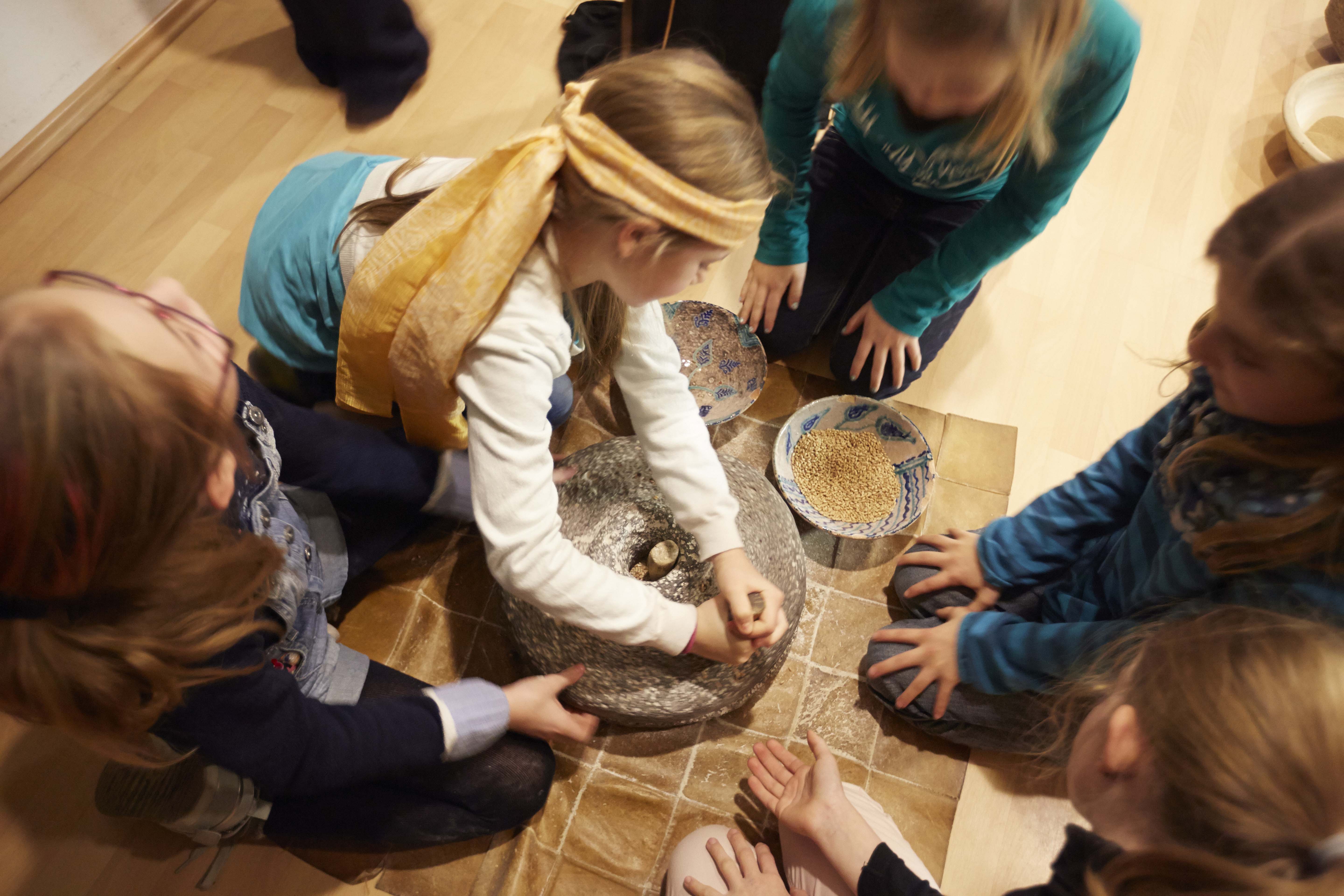 Kinder knien im Kreis um einen Mahlstein und Schüsseln mit verschiedenem Getreide und dürfen mahlen