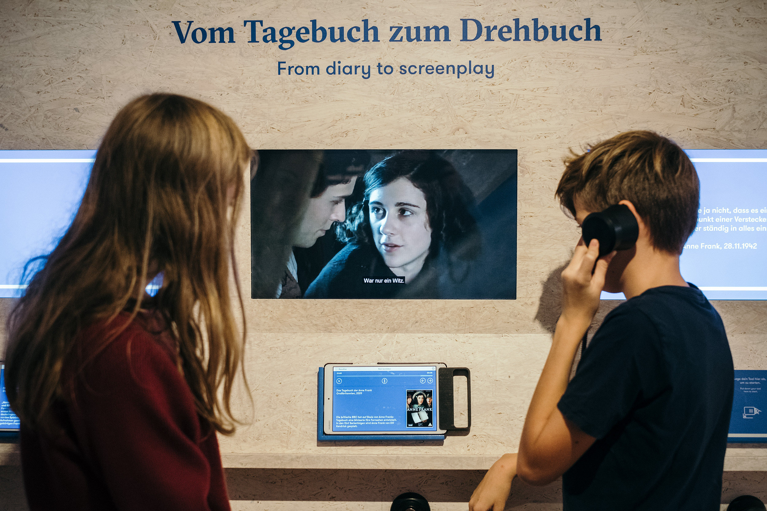 Foto zweier Kinder vor einem Bildschirm, auf welchem ein Film über Anne Frank läuft. Über dem Bildschirm steht "Vom Tagebuch zum Drehbuch. From Diary to Screenplay"